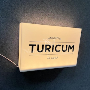 May include: A rectangular, illuminated sign with the word "TURICUM" in bold black letters. The sign also includes the text "HANDCRAFTED" and "IN ZURICH" in a smaller font. A thin wire extends from the sign.