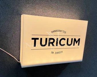 May include: A rectangular, illuminated sign with the word "TURICUM" in bold black letters. The sign also includes the text "HANDCRAFTED" and "IN ZURICH" in a smaller font. A thin wire extends from the sign.