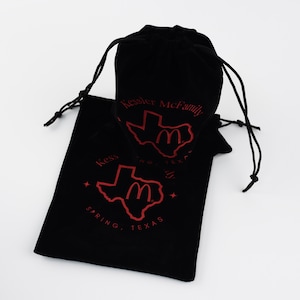 May include: Two black velvet drawstring bags. Each bag has red text and graphics, including a Texas outline with a stylized "M" inside, and the words "Kessler McFamily" and "Spring, Texas."