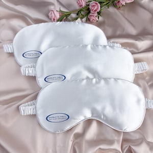 May include: Three white satin sleep masks with elastic bands are arranged on a satin surface. Each mask has a rounded shape and a small, oval logo that reads "BARKATE | NEHI ORAL SLEEP APPLIANCES". Pink roses are in the background.