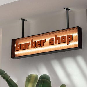 Puede incluir: Un letrero rectangular de barbería con las palabras "barber shop" en letras naranjas. El letrero está suspendido del techo por dos varillas negras. El letrero tiene un brillo cálido y anaranjado.