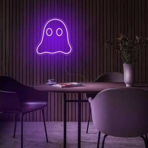 May include: A purple neon ghost sign is mounted on a wooden wall. The ghost has two round eyes and a wavy bottom. The sign is lit up, casting a purple glow. The sign is above a table with chairs.