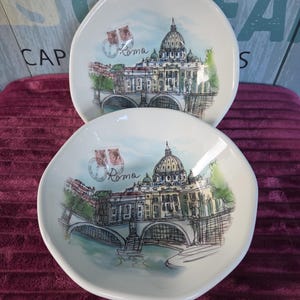 Może przedstawiać: Dwie białe miski ceramiczne z akwarelową ilustracją Bazyliki Świętego Piotra w Rzymie we Włoszech. Wzór zawiera most, rzekę oraz grafikę znaczka pocztowego z napisem Roma wydrukowanym czarną kursywą.
