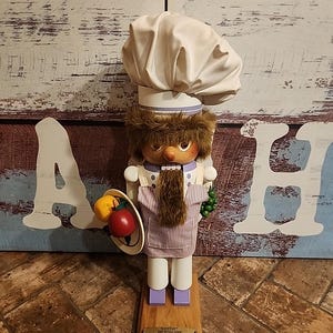 May include: A wooden nutcracker dressed as a chef, wearing a white chef's hat, a purple and white striped apron, and holding a plate with a red apple and a yellow pepper. The nutcracker has a brown beard and brown hair. The nutcracker is standing on a wooden base with a plaque that reads "Westfalia The Original Cook".