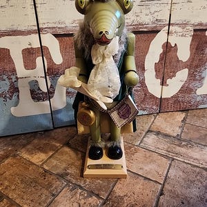 May include: A green and brown wooden nutcracker with a crocodile head, wearing a green jacket, white shirt, and a gold pocket watch. The nutcracker is standing on a wooden base with the text "Peter Pan Series - Crocodile" on it.