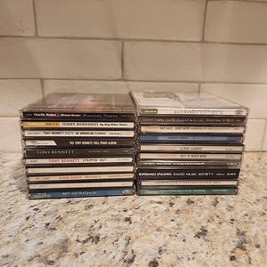 Puede incluir: Dos pilas de CD, cada una en fundas de plástico transparente. Las portadas de los álbumes muestran nombres de artistas y títulos, con géneros como jazz y música vocal. Los CD están colocados sobre una superficie moteada, con una pared de ladrillos al fondo.