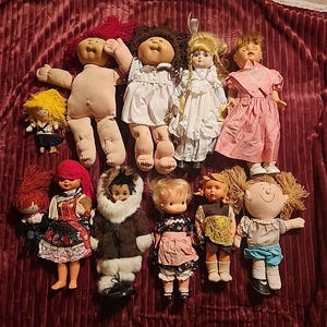 Puede incluir: Una colección de muñecas vintage, incluyendo Cabbage Patch Kids y otras muñecas con diversos peinados y atuendos. Las muñecas están dispuestas sobre una superficie texturizada de color burdeos.