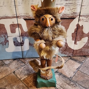 Könnte beinhalten: Eine Holzfigur eines Mäusekönigs, der eine goldene Krone und ein braunes, pelziges Kostüm trägt. Die Maus hat ein Holzgesicht mit schwarzen Augen und einer langen Holz-Nase. Sie steht auf einem grünen Sockel und hält ein rosa Etikett.