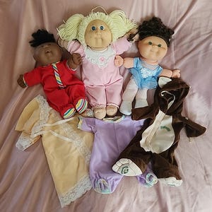 Puede incluir: Cuatro muñecas Cabbage Patch Kids con diferentes atuendos. Una muñeca lleva un pijama a rayas rojas y blancas, otra lleva un pijama rosa, otra lleva un atuendo de bailarina azul y blanco, y la última muñeca lleva un disfraz de oso marrón.