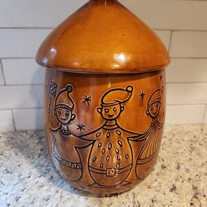 Pode incluir: Um pote de cerâmica castanho com tampa a condizer, apresentando um design em relevo de três figuras com gorros pontiagudos de mãos dadas. As figuras estão rodeadas por pequenos detalhes em forma de estrela e a peça tem um acabamento brilhante.