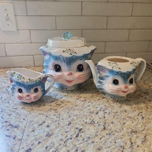 May include: A ceramic kitchen set featuring a creamer, a lidded canister, and a teapot, all shaped like cat faces. Each piece is painted with blue and white fur details, pink noses, and small blue floral accents on the white rims and lids.