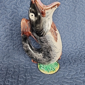 May include: A ceramic fish-shaped jug, predominantly grey with brown and pink details, and a green base with yellow edging. The open-mouthed fish jug sits on a blue quilted surface. The jug is a decorative item.