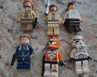 May include: Six Lego Star Wars minifigures are displayed. The figures include characters with tan, white, blue, and orange uniforms. One figure holds a blue lightsaber. Each figure has a black blaster pistol. The figures are on a speckled surface.