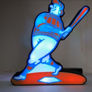 May include: A lighted baseball player figurine in a batting stance. The figure is outlined in blue with orange accents, holding a bat. The base is orange, and the figure is illuminated with a soft glow, creating a vintage aesthetic.
