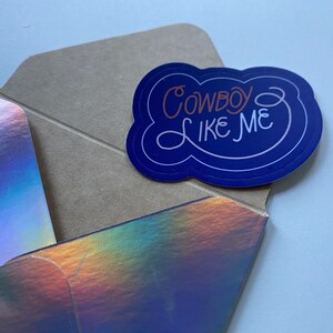 Pode incluir: Um adesivo azul e branco com o texto "Cowboy Like Me" em uma fonte branca cursiva. O adesivo est&aacute; em um envelope marrom com um acabamento hologr&aacute;fico iridescente.