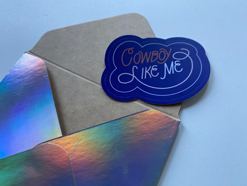 Pode incluir: Um adesivo azul e branco com o texto "Cowboy Like Me" em uma fonte branca cursiva. O adesivo est&aacute; em um envelope marrom com um acabamento hologr&aacute;fico iridescente.