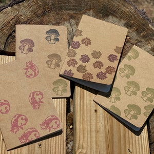 May include: Four small kraft paper notebooks with different nature-themed designs. One notebook has a pattern of mushrooms, another has a pattern of leaves, another has a pattern of ladybugs, and the last one has a pattern of mushrooms.