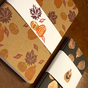 May include: Two notebooks with fall-themed covers. The larger notebook has a brown cover with a white band and a pattern of leaves and pumpkins. The smaller notebook has a black cover with a gold pattern of pumpkins and a white band with a jack-o'-lantern.