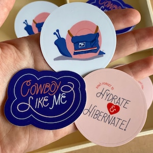 Pode incluir: Um conjunto de quatro adesivos com designs diferentes. Um adesivo &eacute; azul e apresenta um caracol de desenho animado vestindo um uniforme de carteiro azul e rosa. Outro adesivo &eacute; rosa e apresenta o texto "N&atilde;o se esque&ccedil;a de se hidratar e hibernar!" em branco com um cora&ccedil;&atilde;o vermelho. O terceiro adesivo &eacute; azul e apresenta o texto "Cowboy Like Me" em branco. O quarto adesivo &eacute; branco e apresenta o texto "Cowboy" em azul.