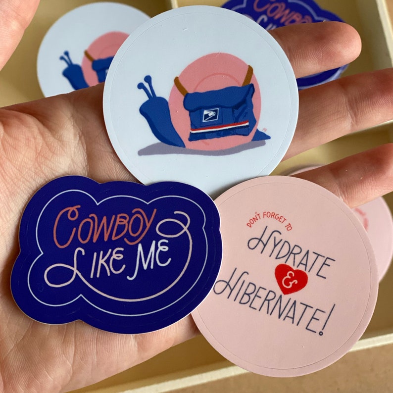 Pode incluir: Um conjunto de quatro adesivos com designs diferentes. Um adesivo &eacute; azul e apresenta um caracol de desenho animado vestindo um uniforme de carteiro azul e rosa. Outro adesivo &eacute; rosa e apresenta o texto "N&atilde;o se esque&ccedil;a de se hidratar e hibernar!" em branco com um cora&ccedil;&atilde;o vermelho. O terceiro adesivo &eacute; azul e apresenta o texto "Cowboy Like Me" em branco. O quarto adesivo &eacute; branco e apresenta o texto "Cowboy" em azul.