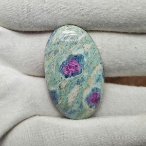 May include: An oval gemstone with a light green base, speckled with white and blue. It has three magenta-coloured inclusions. The polished stone is set against a white fabric background.
