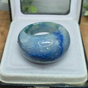 Puede incluir: Un anillo ancho y redondo hecho de piedra azul y verde. El anillo tiene un aspecto veteado con vetas de azul más oscuro y verde más claro. Se exhibe en una caja de joyería negra con interior blanco. Diámetro aproximado de 2,5 cm.