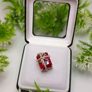 May include: A silver-toned pendant in the shape of a gift box, featuring a large rectangular red gemstone. The pendant is adorned with a bow and small clear gemstones. It rests inside a black jewelry box with a white interior.
