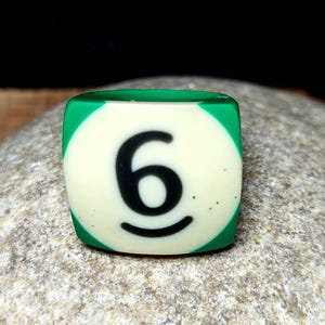 May include: A square-shaped ring featuring a green border and a central cream-colored circle with the black number 6 printed in the middle. The design mimics the appearance of a billiard ball, with smooth surfaces and rounded edges on the band.