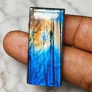 Könnte beinhalten: Ein rechteckiger Labradorit-Edelstein-Cabochon mit polierter Oberfläche und einem lebendigen, irisierenden blauen und goldgelben Schimmer. Der Stein weist natürliche interne Risse und vertikale Streifen in seiner gesamten Struktur auf.