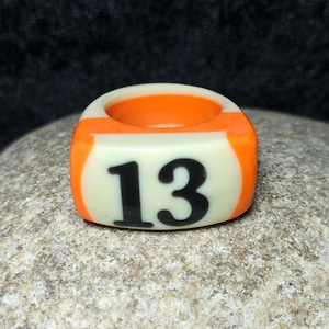 Può includere: Un anello massiccio caratterizzato da una superficie liscia color bianco sporco con il numero 13 nero stampato sul davanti. L'anello presenta una fascia spessa e arrotondata con vivaci accenti arancioni sui lati e sulla parte superiore.
