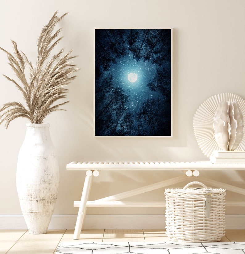 May include: A white bench with a white wicker basket and a white vase with dried pampas grass. A framed print of a full moon in a dark blue forest hangs on the wall behind the bench.