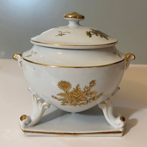 May include: A white porcelain serving dish with a lid, adorned with gold floral patterns and trim. The dish has a rounded body, two small handles, and sits on a rectangular base with ornate legs. The lid is topped with a gold knob.