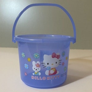 Puede incluir: Un cubo de plástico azul con asa que presenta a Hello Kitty y un conejito. El cubo tiene un diseño floral y el texto "HELLO KITTY".