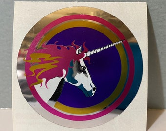 Puede incluir: Una pegatina circular plateada con un diseño rosa, dorado y morado. En el centro aparece un unicornio blanco con una crin rosa y un cuerno dorado.