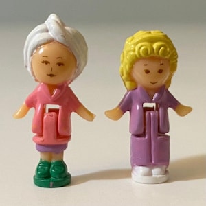 May include: Two small plastic figures. One wears a pink robe, a white towel on their head, and green shoes. The other wears a purple robe, has yellow hair, and white shoes. Both figures have their arms outstretched.