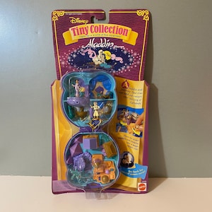 May include: A blue and purple plastic toy playset featuring Aladdin and Jasmine from the Disney movie Aladdin. The playset is a miniature version of the palace in the movie and includes a magic carpet, a treasure chest, and other accessories. The playset is packaged in a clear plastic box with a colorful cardboard backing.