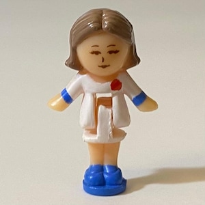 May include: A small plastic figure of a person in a white coat with blue details and blue shoes. The figure has brown hair and a red flower on the coat. The figure is standing with arms outstretched.