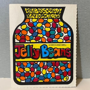 Puede incluir: Una colorida pegatina con un tarro lleno de gominolas. El tarro está delineado en negro, con una parte superior amarilla y las palabras "Jelly Beans" en rojo, azul y amarillo. La pegatina tiene las palabras "SCRATCH AND SMELL" encima del texto.