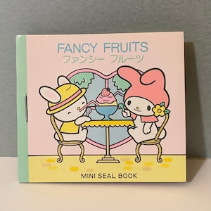 Puede incluir: Un mini libro de sellos con una portada rosa con el texto "FANCY FRUITS" en inglés y japonés. La ilustración muestra dos personajes de dibujos animados disfrutando de un helado. El libro tiene una lomo verde y un borde amarillo.