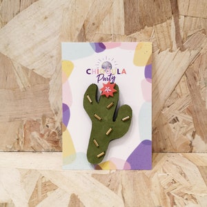 Peut inclure: Une épingle en forme de cactus vert avec des détails dorés et une fleur rouge, présentée sur une carte avec le texte "CHICOGULA PARTY". La carte présente un design abstrait coloré. L'épingle est probablement un accessoire décoratif.