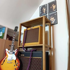 Può includere: Una mensola in legno con un amplificatore per chitarra giallo e marrone e un amplificatore VOX nero con un motivo a rombi marroni. Una chitarra elettrica sunburst è a sinistra. Quattro ritratti incorniciati dei Beatles sono appesi al muro.
