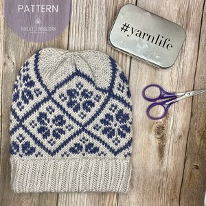 May include: A knitted hat with a geometric pattern in shades of blue and white. The hat is on a wooden surface with a metal tin that says #yarnlife.