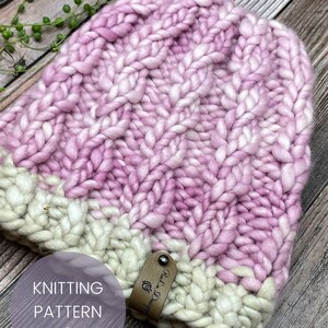 May include: A pink and white knitted hat with a cable stitch pattern. The hat has a leather tag with the text "Rely on D's" on it. The hat is on a wooden surface.