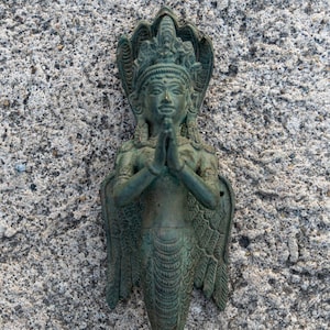 Può includere: Una statua di metallo verde di una divinità con una corona a forma di serpente e ali, con le mani giunte in posizione di preghiera. La statua è in piedi su una superficie testurizzata.