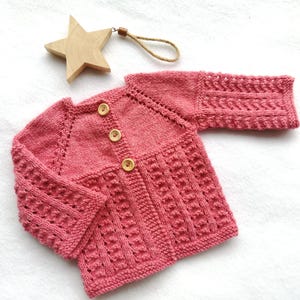 Op de afbeelding: Een handgemaakt, roze babyvestje met houten knopen. Het gebreide vest heeft textuurpatronen op het lijfje en de mouwen. Een houten ster ornament ligt ernaast.