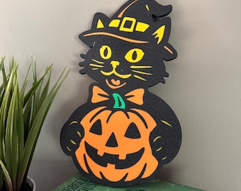 May include: A black cat cutout wearing a yellow-banded witch hat and an orange bow tie, holding an orange jack-o'-lantern with a carved face. The cat has bright yellow eyes and a small mouth, rendered in a flat, graphic style with bold outlines.