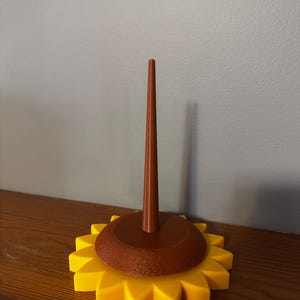 May include: A yellow and brown sunflower-shaped yarn holder. The base is a bright yellow with pointed petals. A brown, circular center supports a tall, brown spindle for holding yarn. The yarn holder is on a wooden surface.