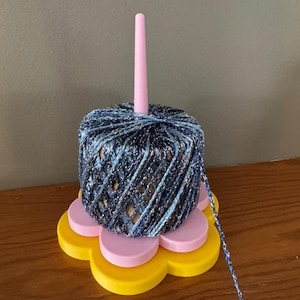 May include: A yarn holder with a blue and silver yarn ball. The yarn ball sits on a pink and yellow flower-shaped base. A pink spindle rises from the center of the yarn ball. The yarn is a mix of blue and silver.