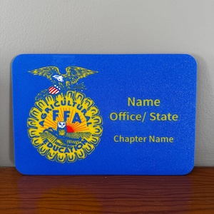 May include: A blue name tag with rounded corners. The left side features a yellow and blue emblem with an eagle and the letters "FFA". The right side has text fields for "Name", "Office/ State", and "Chapter Name".