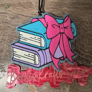 May include: A hanging ornament featuring a stack of two books, one blue and one purple, tied with a large bright pink bow. Below the books is a pink marbled acrylic nameplate with the text Korals Krafts Shop in a cursive white font.